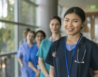 A female doctor of Asian descent poses with her multi-ethnic team of doctors and nurses and smiles directly at the camera while standing in a hospital corridor.