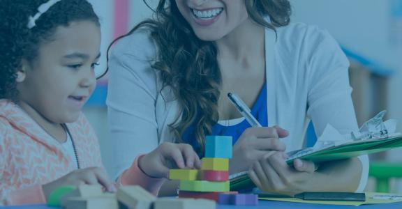 Teacher smiling and taking notes while young girl stacks col